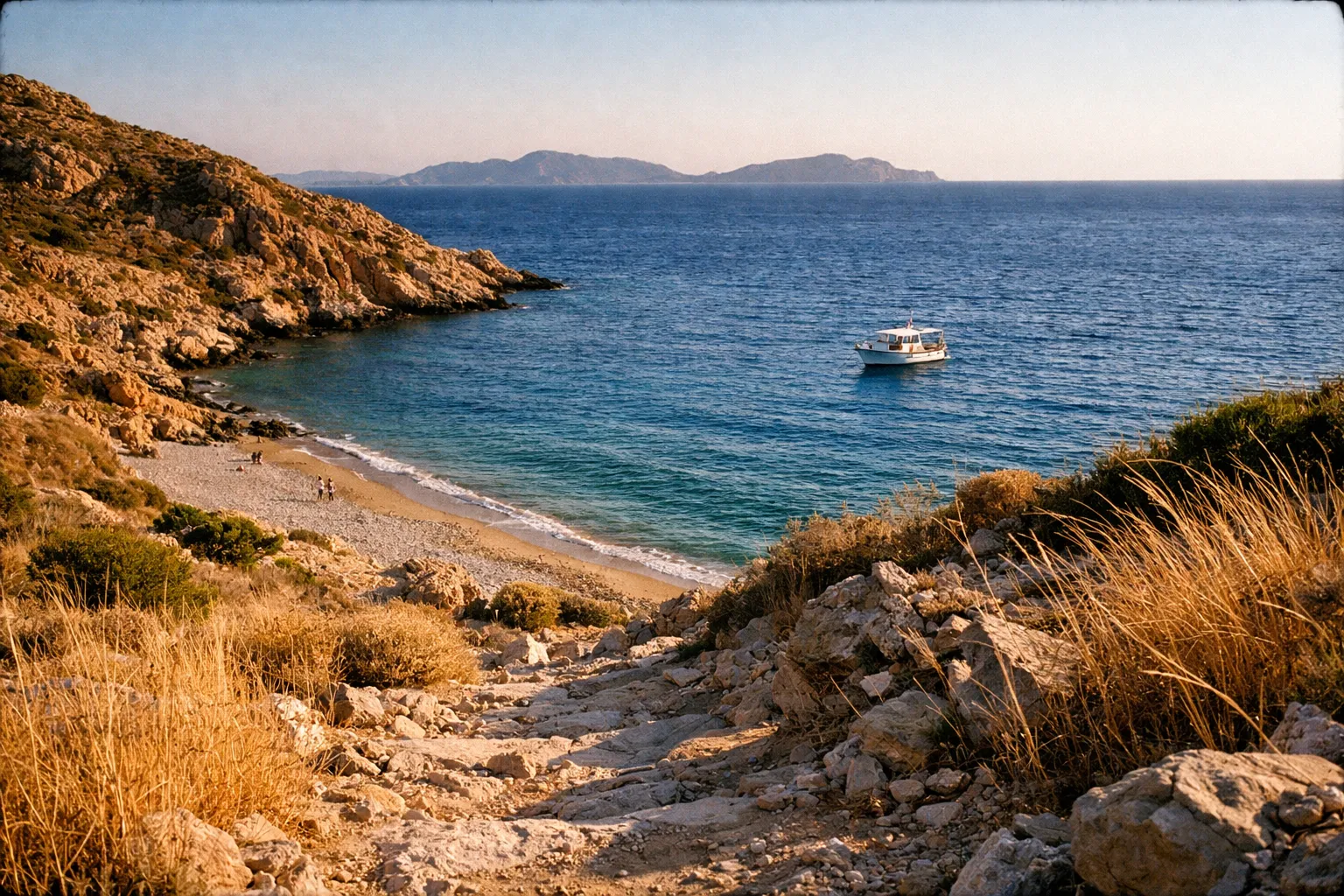 A rocky path leads to a small beach with a boat anchored in the blue sea near Patmos, Greece.