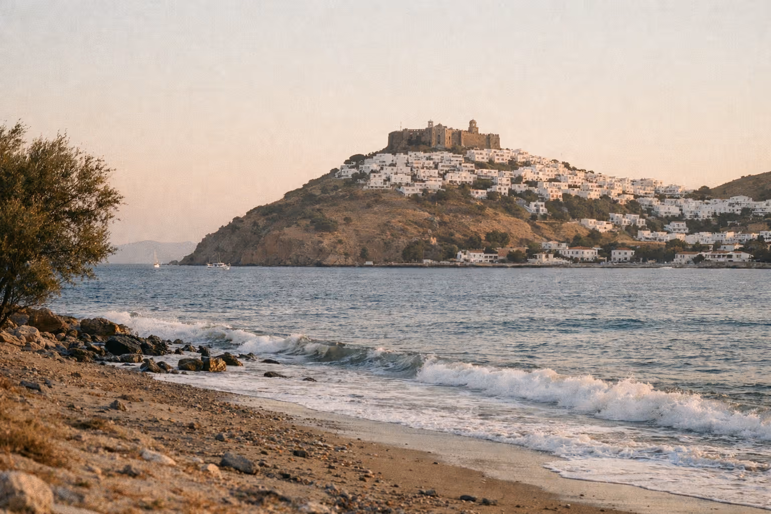 The Monastery of St. John stands atop a hill above white houses in Chora, Patmos, viewed from a pebbly beach.