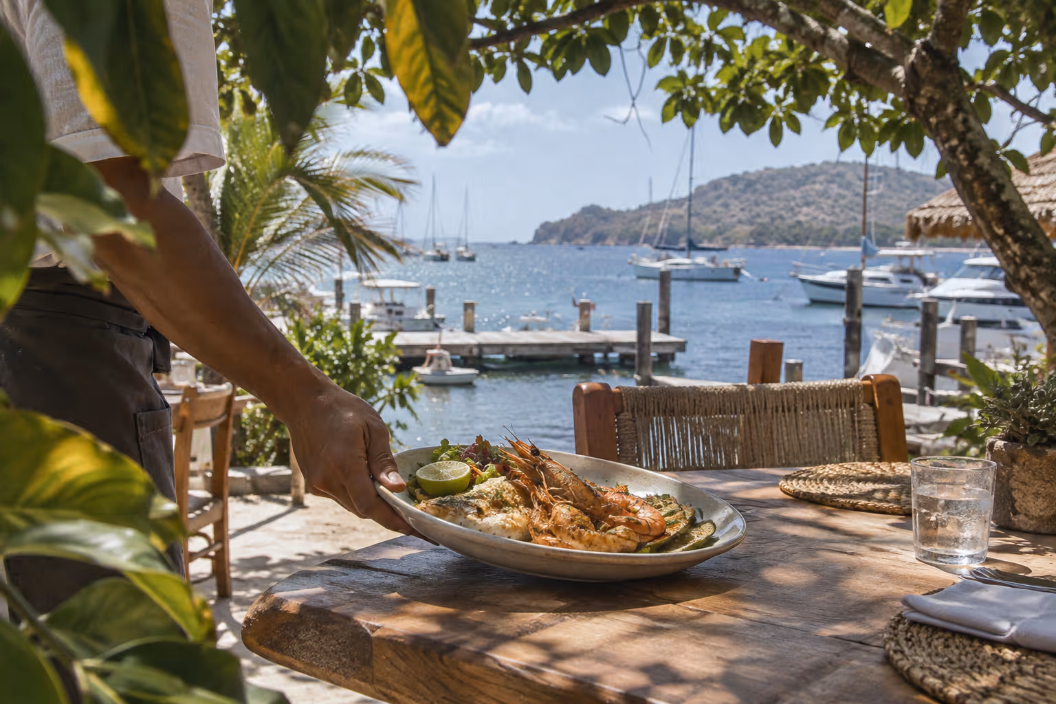 A server brings a seafood platter to a shaded waterfront table overlooking a boat-filled bay.