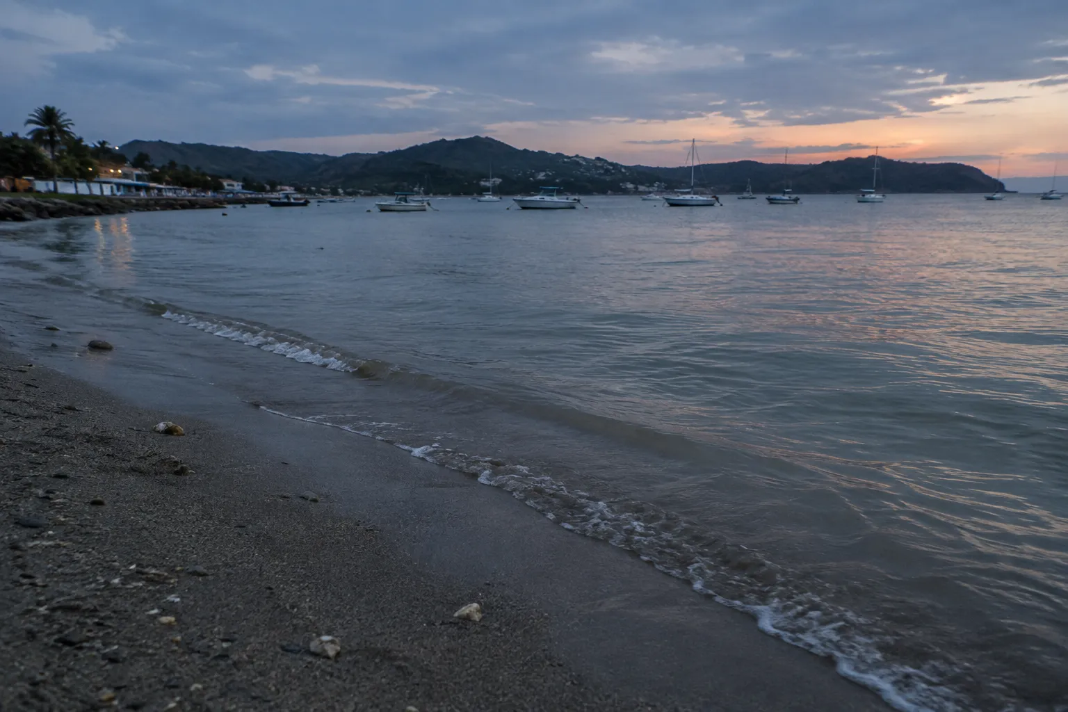 Boats anchor in a coastal bay beside a pebble beach at sunset.