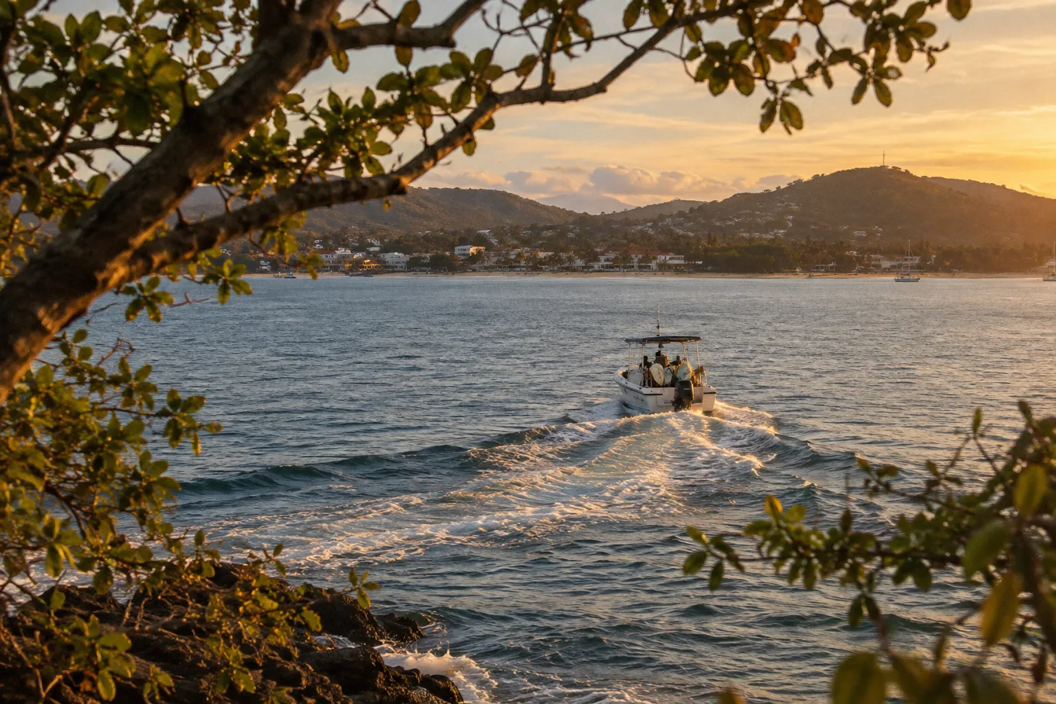A small motorboat crosses a coastal bay near a hillside town at sunset.