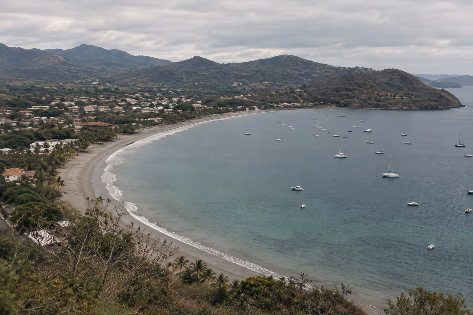 A curved beach and coastal town border a bay dotted with boats beneath dry hills.