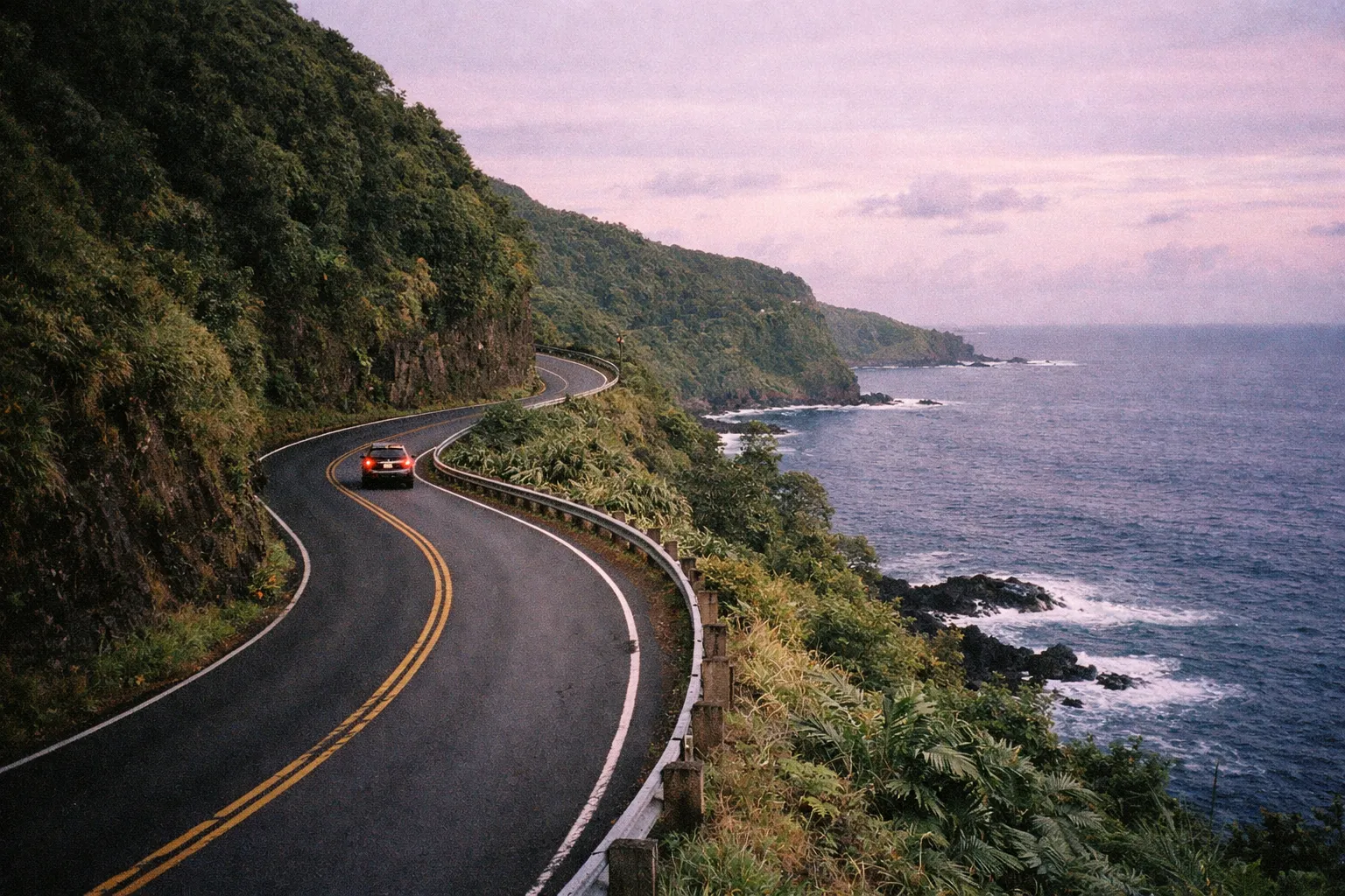 A car drives along the winding Road to Hana next to lush cliffs and the Pacific Ocean.