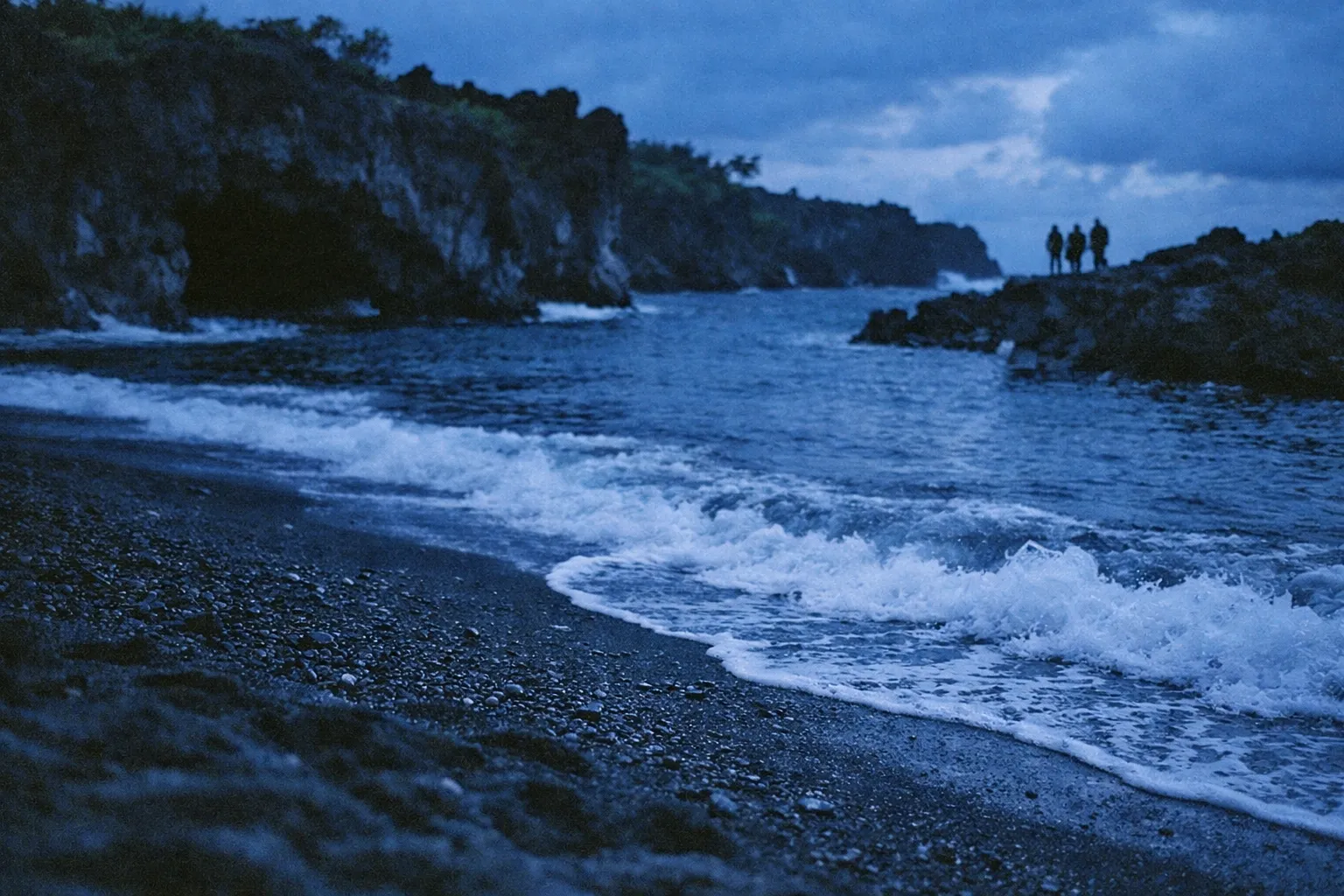 Waves wash onto a rocky black sand beach with cliffs and three people standing on a distant outcrop.