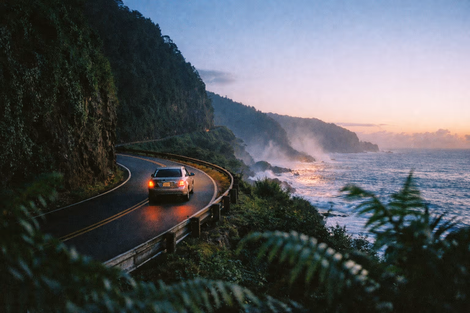 A car drives along a winding coastal road beside cliffs and the ocean at sunset.