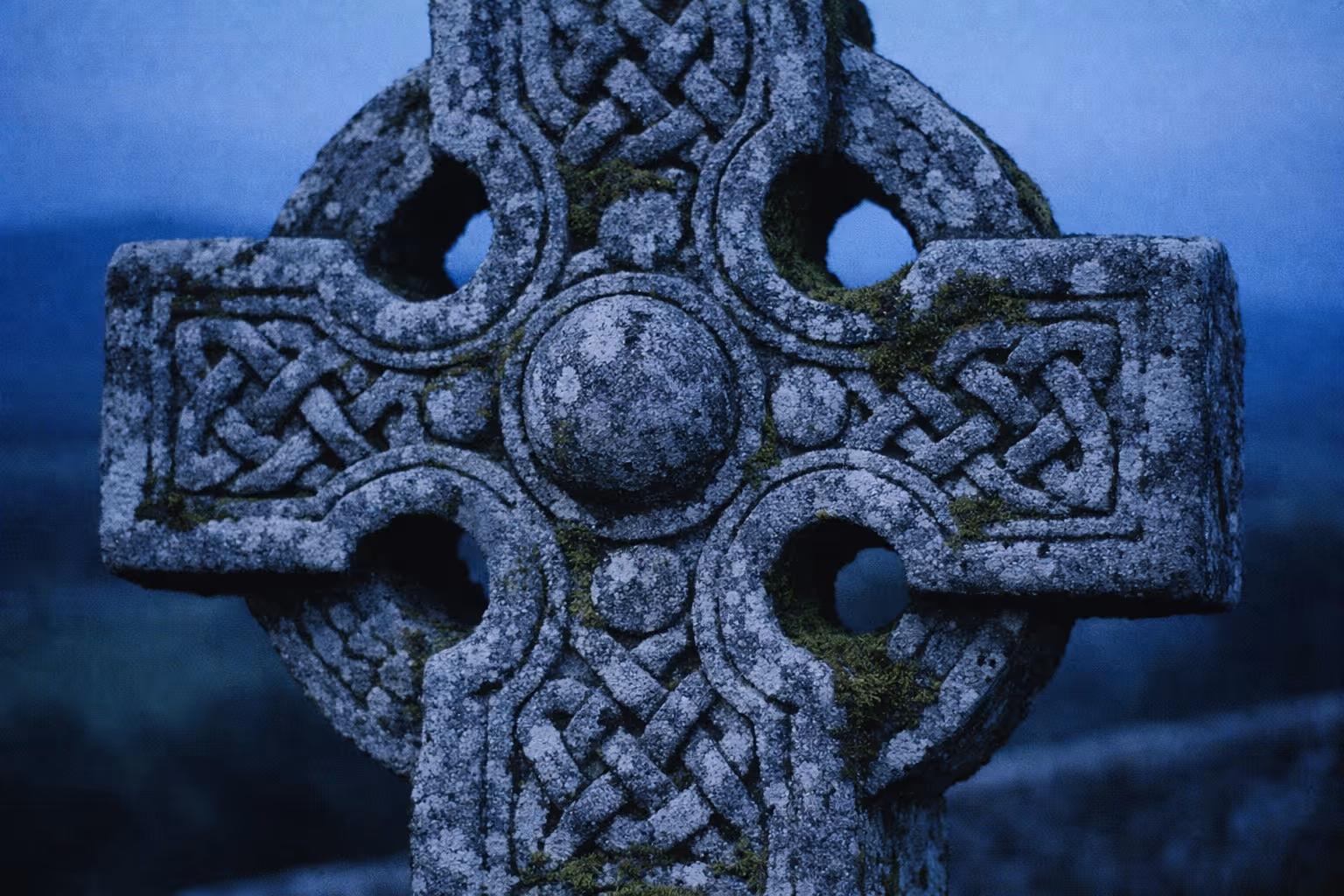 Close-up of a weathered Celtic cross covered in moss against a blue twilight sky.