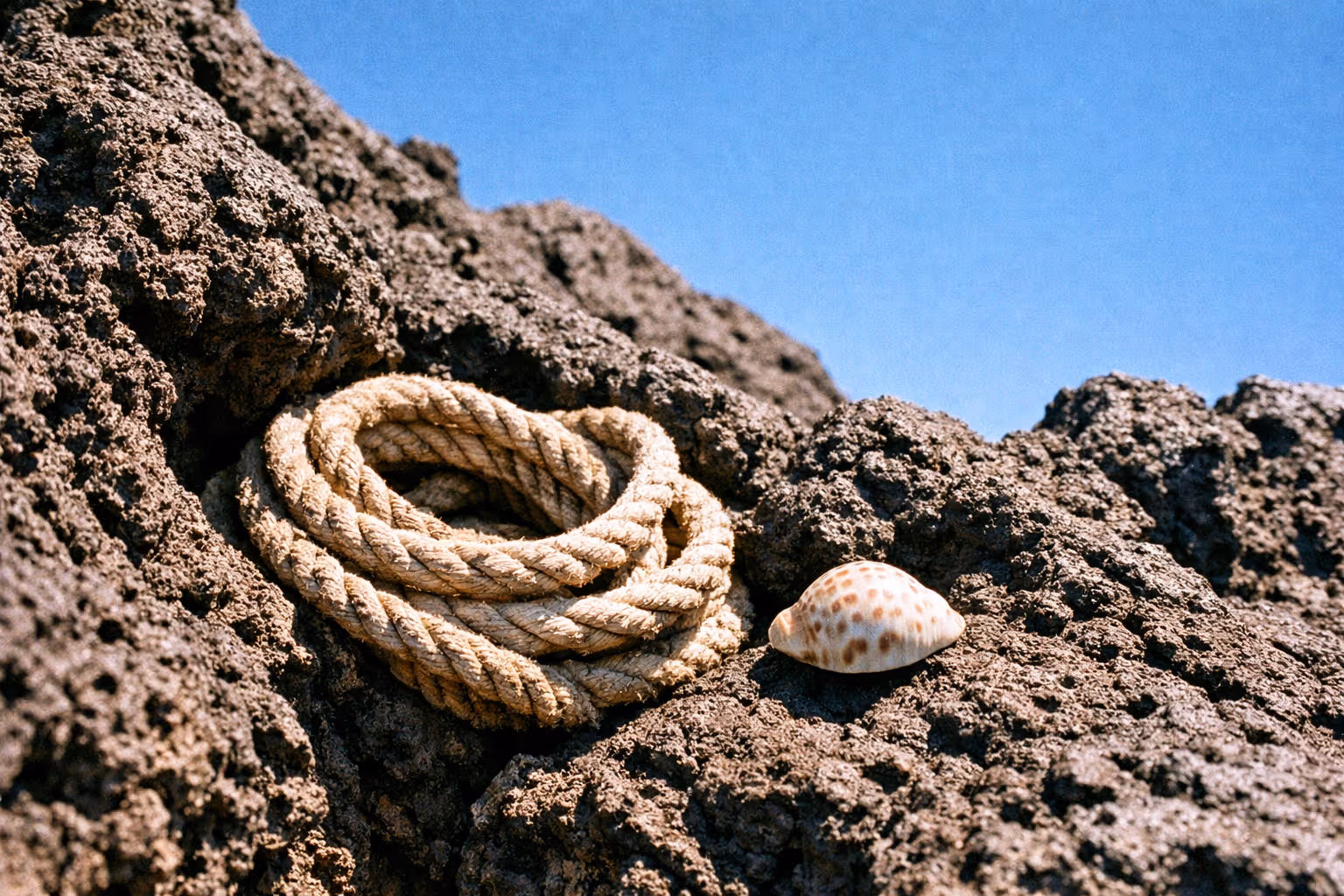 A coil of rope and a seashell rest on volcanic rocks under a clear blue sky.