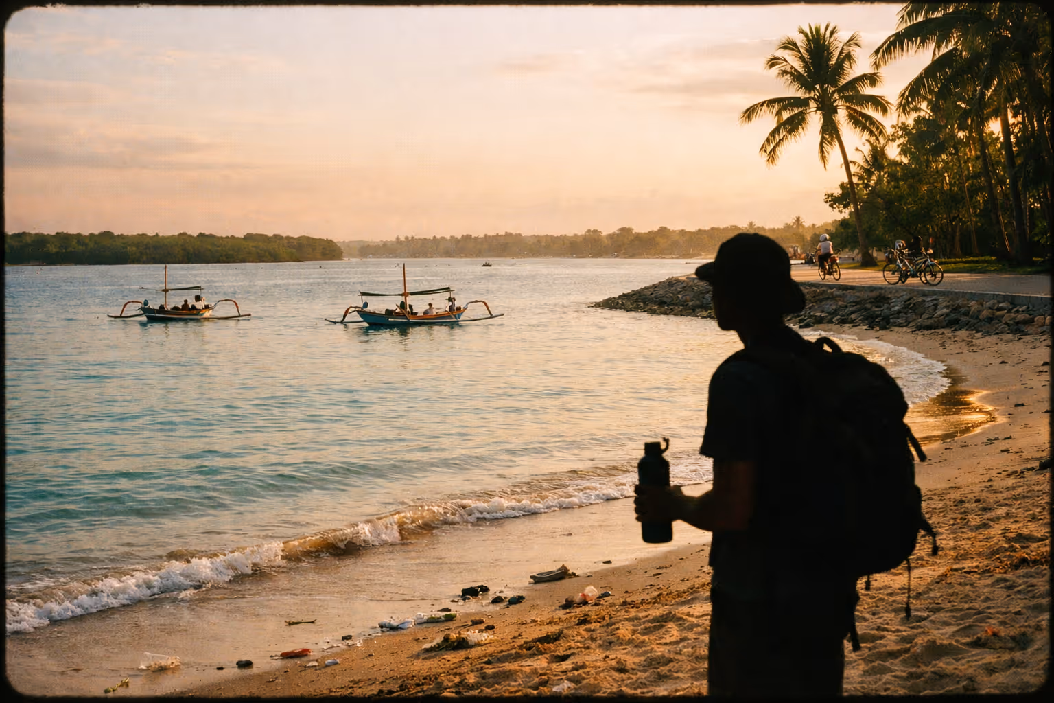 Sanur Beach at sunrise: calm water, a resort-fronted shoreline, distant mangroves and debris along the high-tide line.
