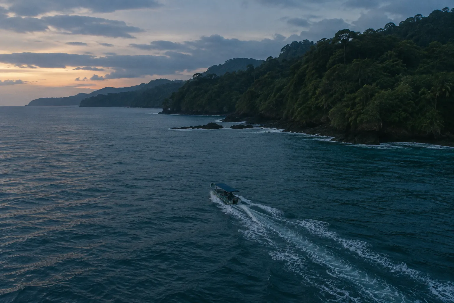 A small boat crosses coastal waters beside a forested shoreline at sunset.