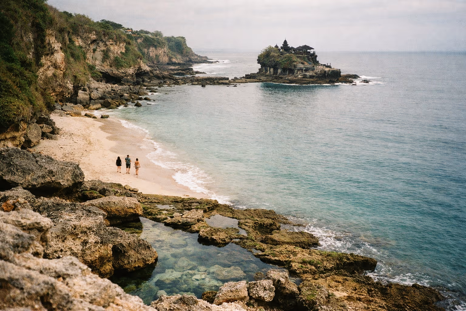Three people walk along a rocky beach with a coastal temple visible on an offshore rock in Bali.