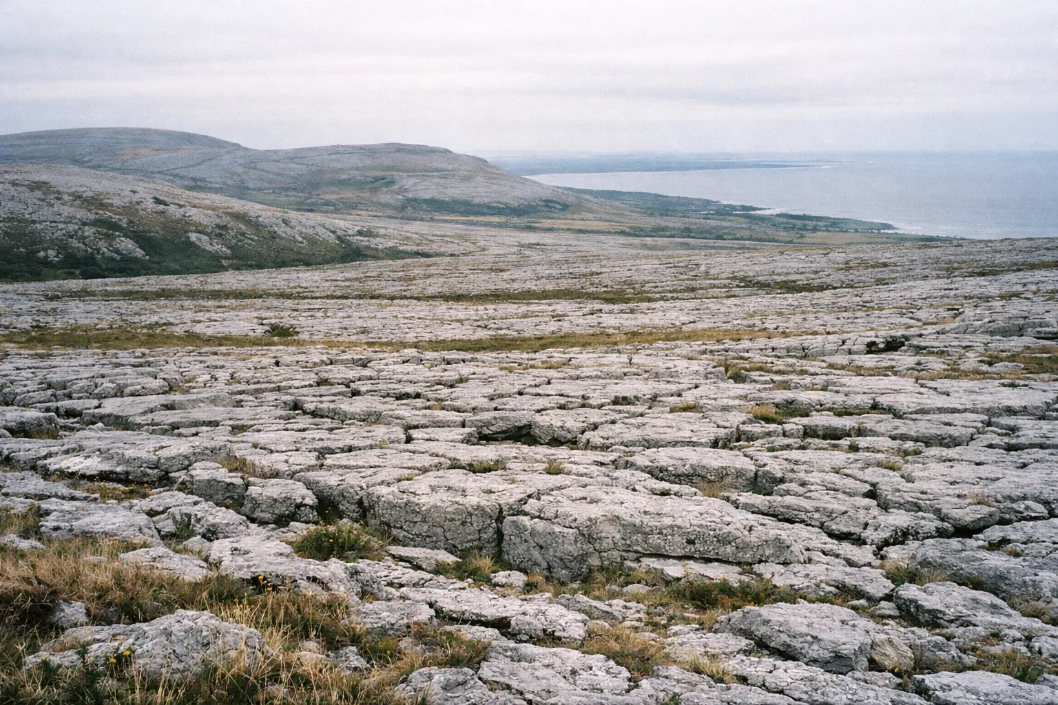Limestone pavement fields of the Burren stretch toward distant hills and the coastline under a cloudy sky.