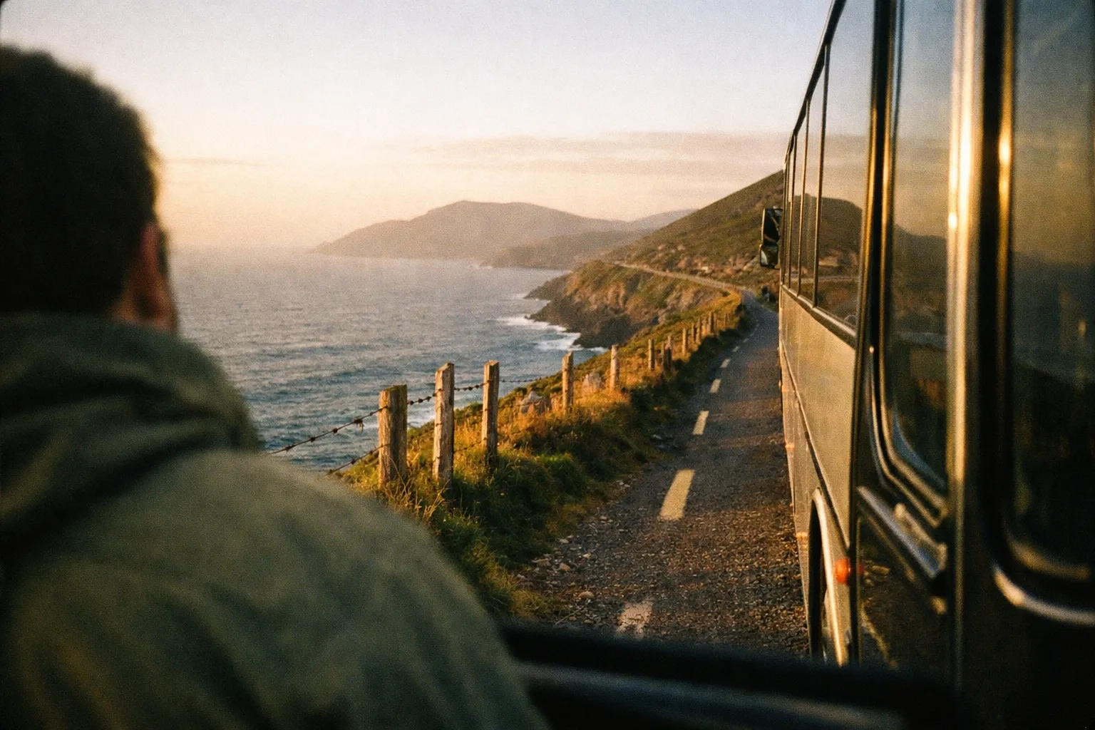 A person looks out from a bus traveling along a coastal road with mountains and sea in view.