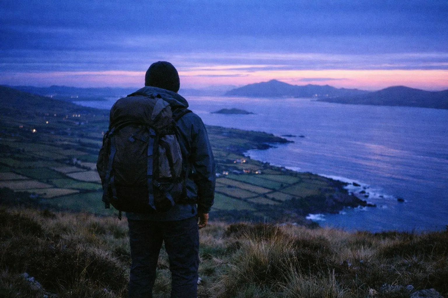 A hiker with a backpack looks out over coastal cliffs and farmland along the Ring of Kerry at sunset.
