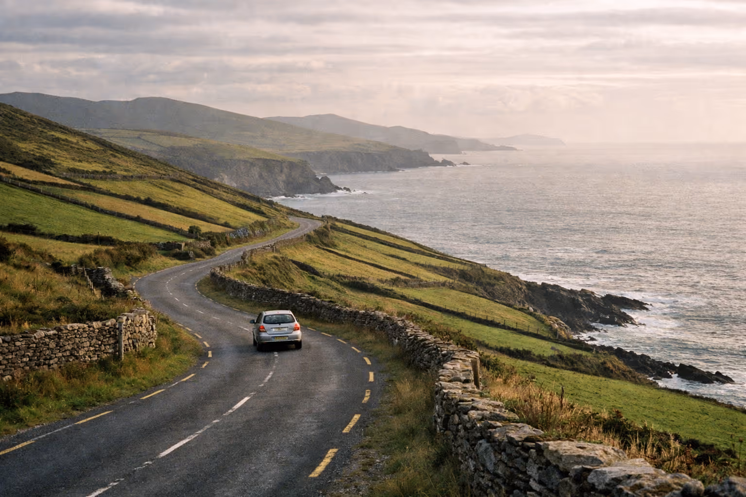A car drives along a winding coastal road overlooking cliffs and the sea on the Ring of Kerry in Ireland.