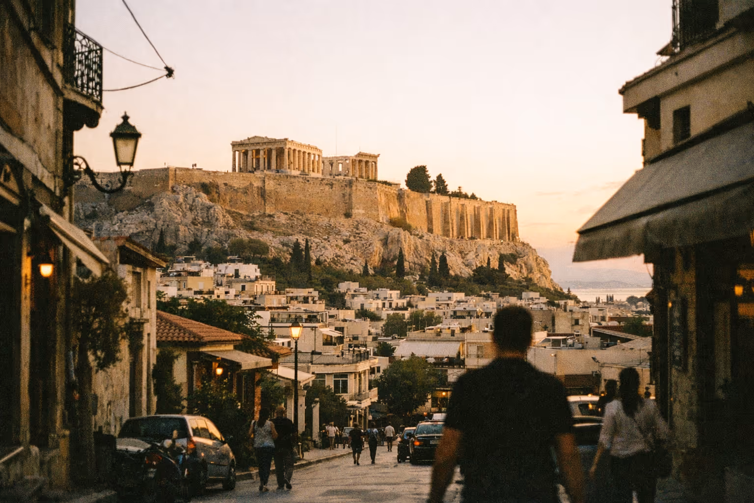 The Acropolis rises above city streets in Athens at sunset as people walk below.