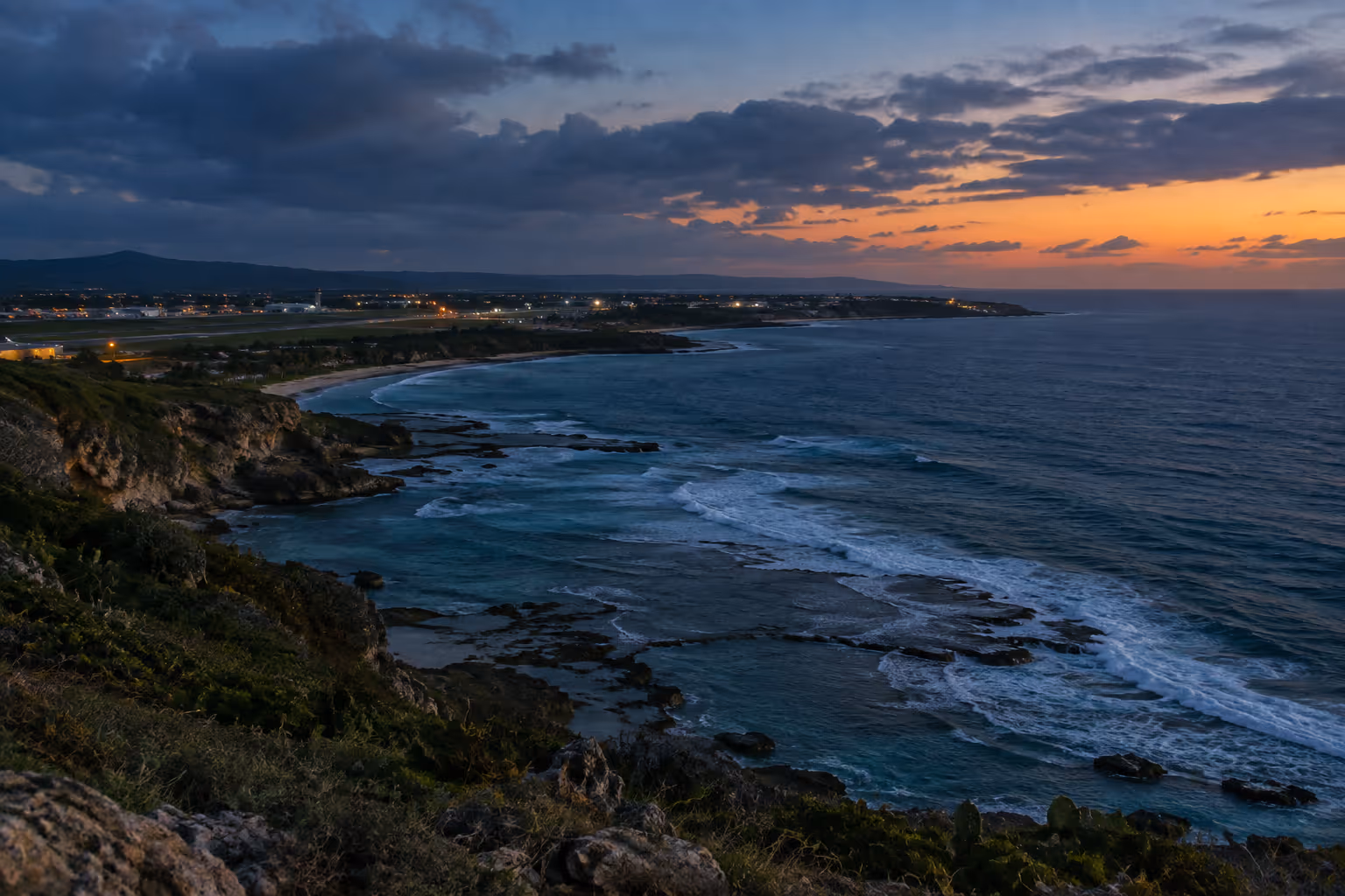 Rocky coastline and surf near Aguadilla, with an airport along the shore at sunset.