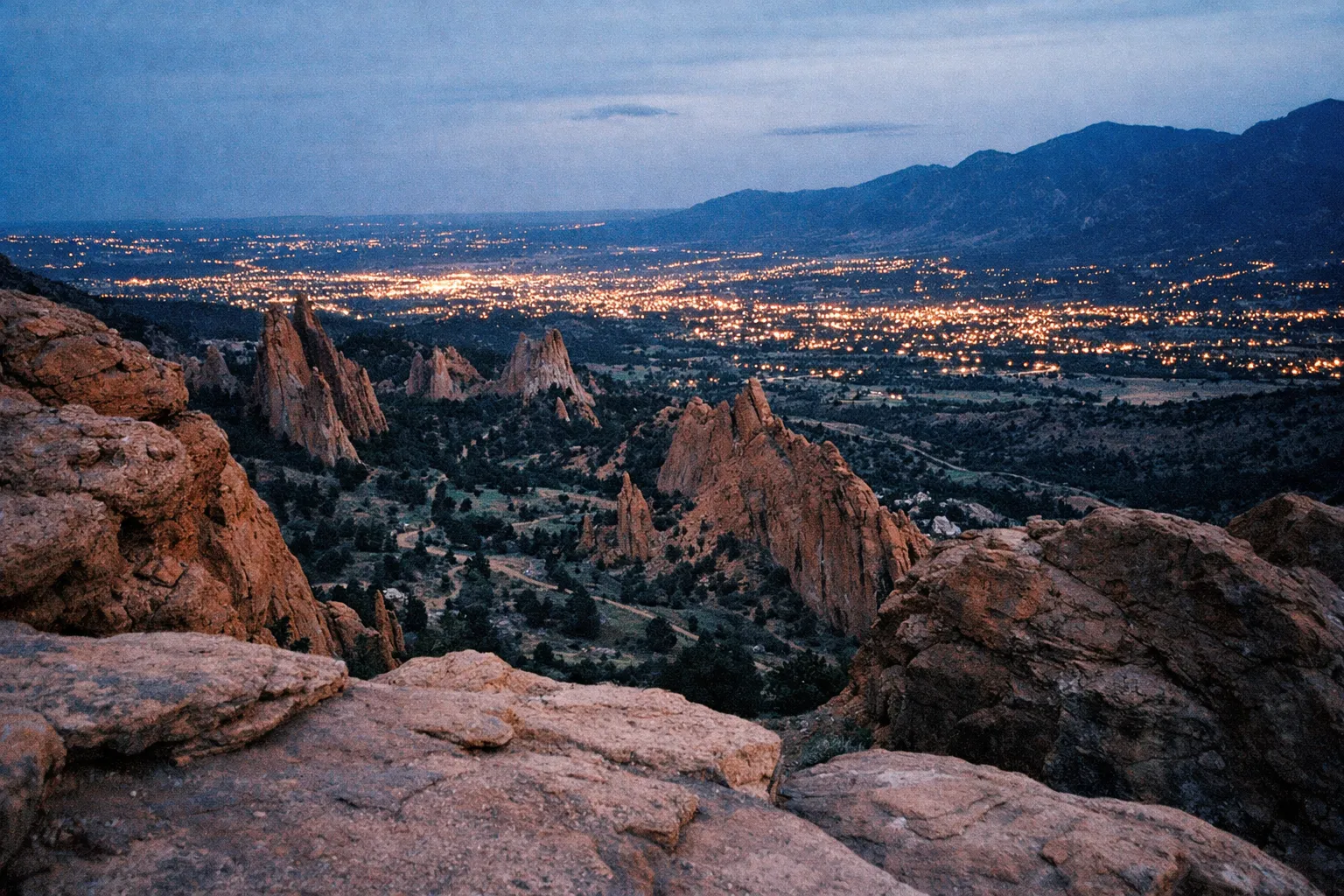 Red rock formations of Garden of the Gods with city lights of Colorado Springs in the distance at dusk.