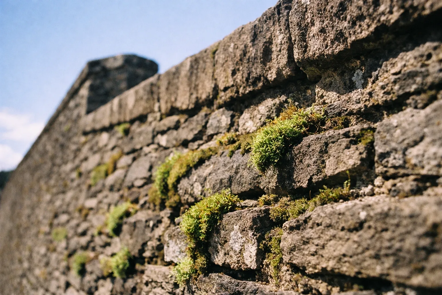 An old stone wall with moss growing between the stones under a blue sky.