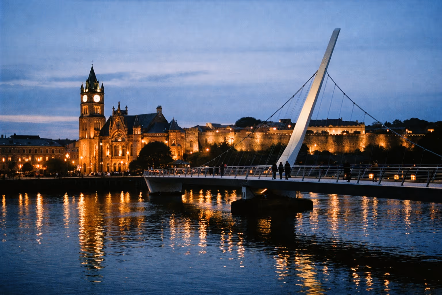 People walk across the Peace Bridge in Derry with the Guildhall and city walls lit up at dusk.