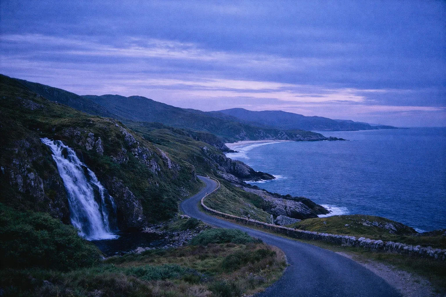 A winding coastal road passes a waterfall and cliffs along the Donegal coastline at dusk.