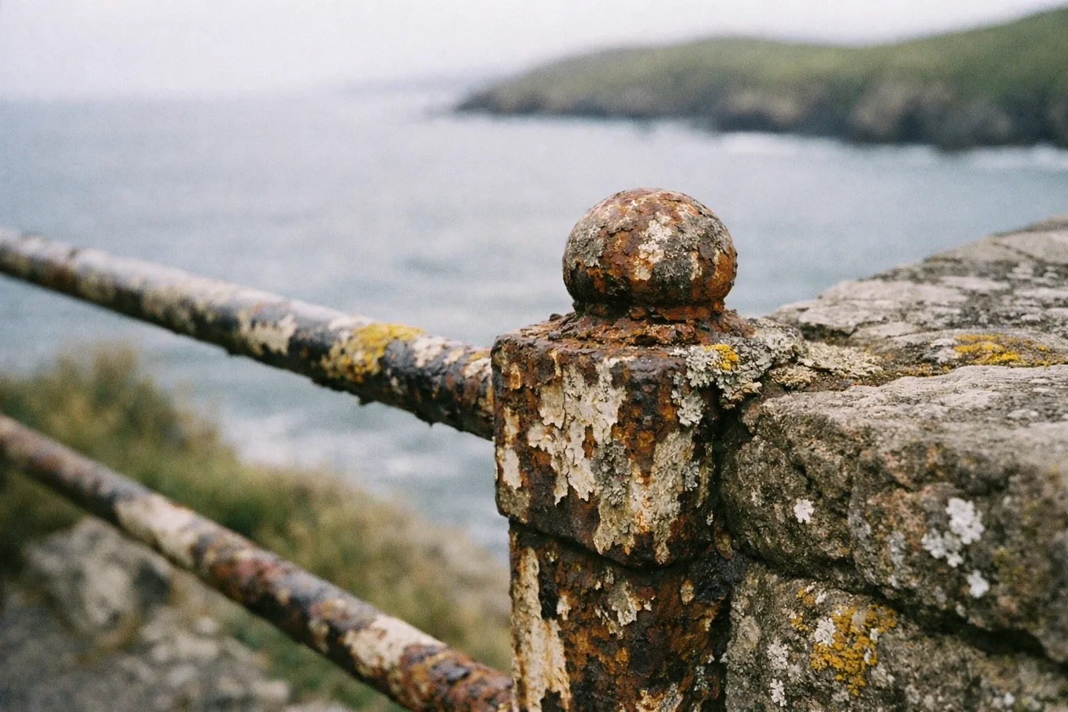 A rusted metal railing overlooks the sea with grassy cliffs in the background.