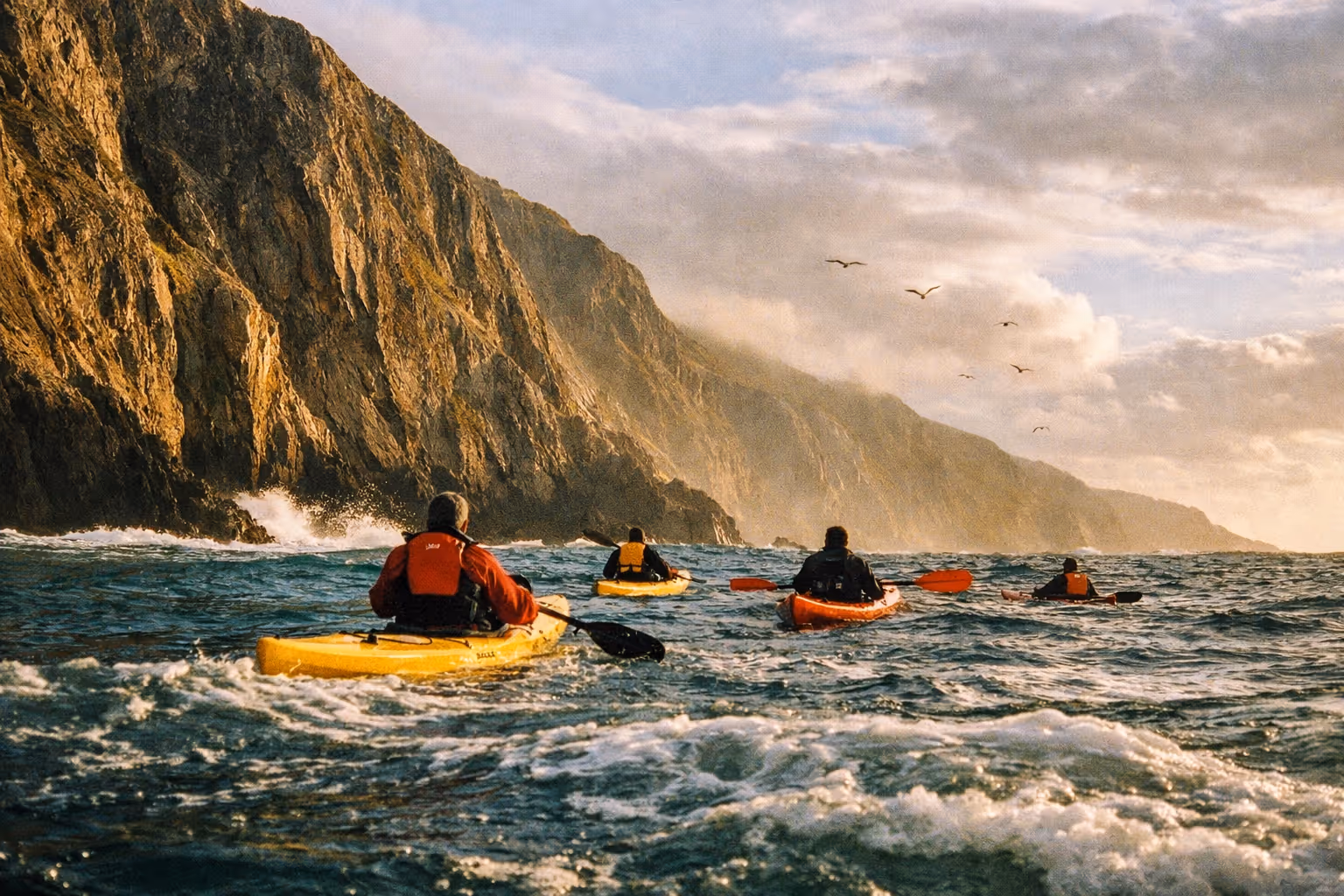 Kayakers paddle near steep sea cliffs along a rugged coastline under a cloudy sky.
