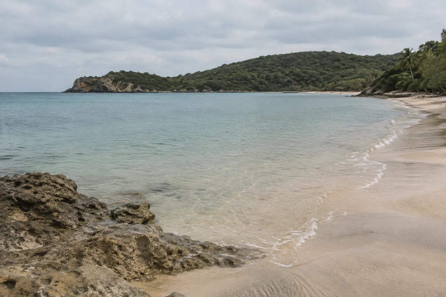 A quiet sandy beach curves around clear water beneath a green, rocky headland.