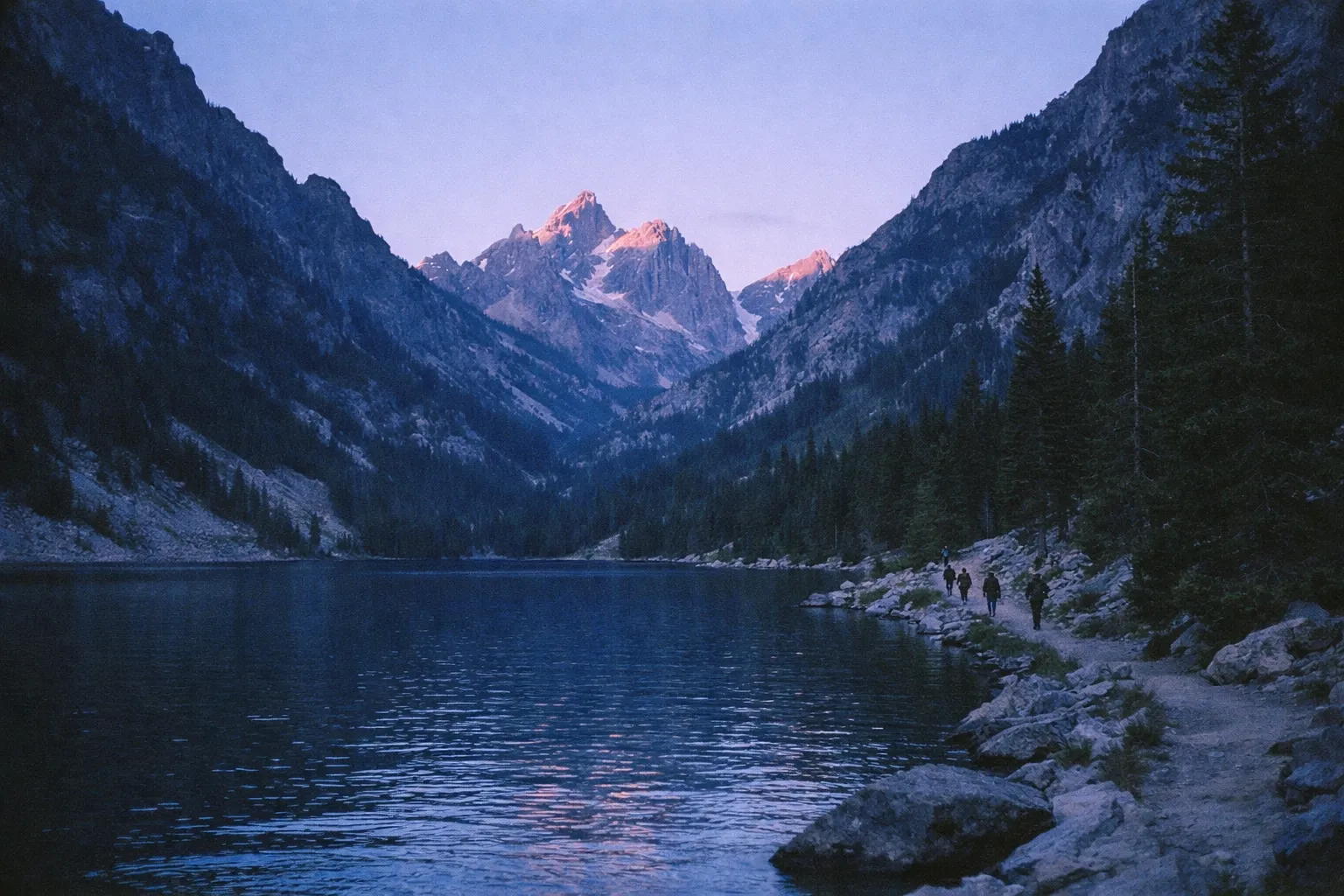 Hikers walk along a trail beside Jenny Lake with the Teton mountains rising in the background.