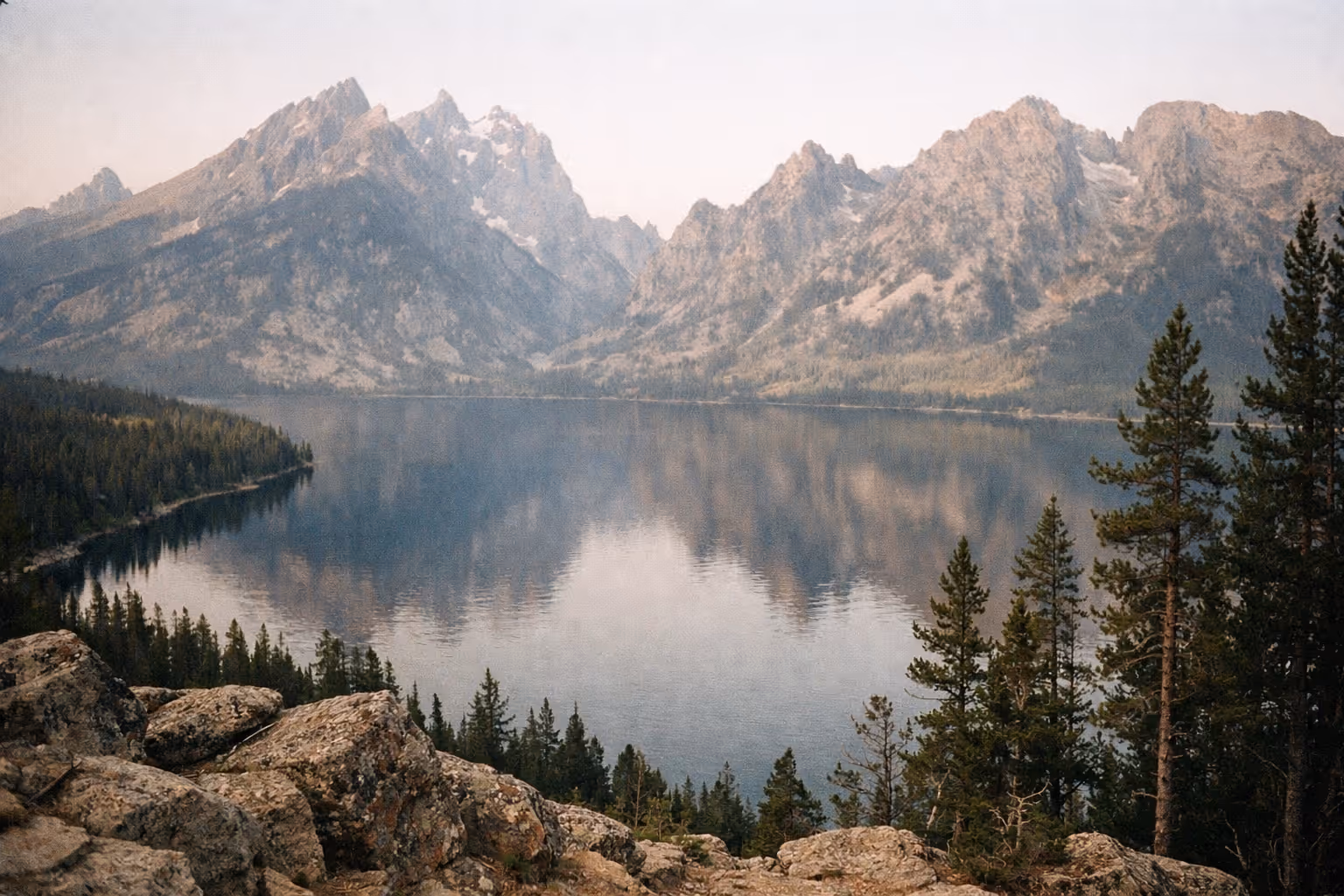 Rocky shoreline and pine trees beside Jenny Lake with the Teton mountain range in the background.