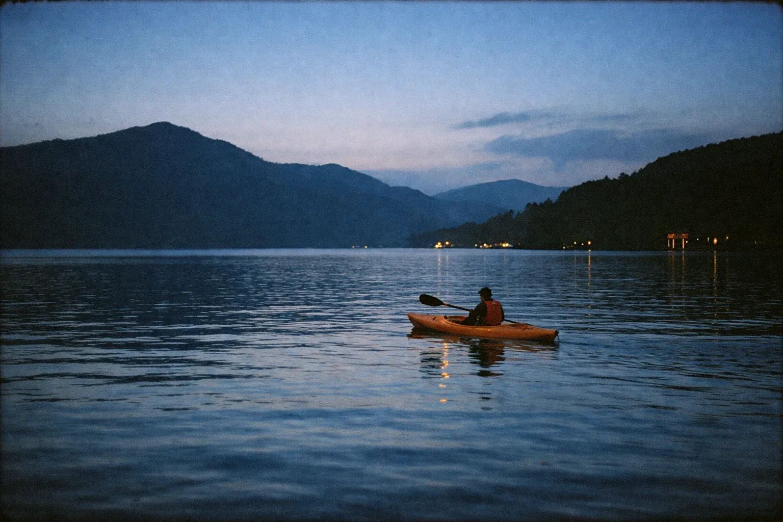 A person paddles a kayak on Lake Ashi at dusk, surrounded by mountains in Hakone.