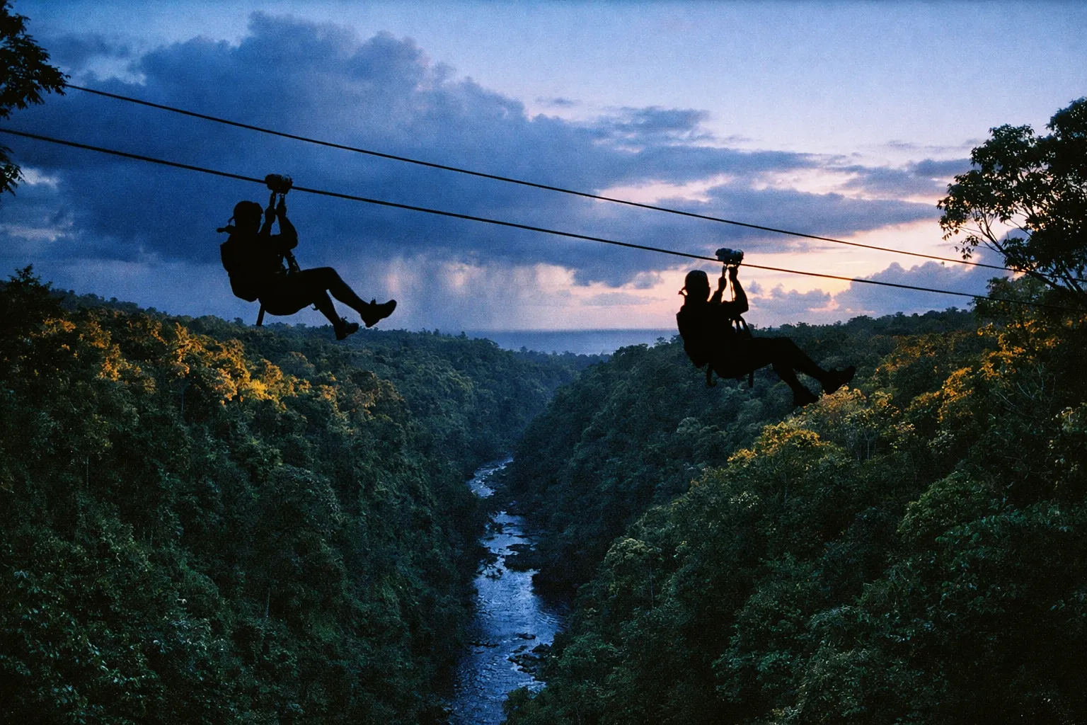Two people zipline high above a lush green valley with a river running below at sunset.