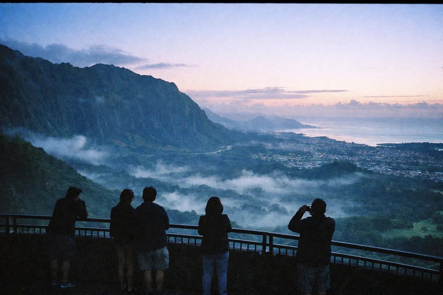 People stand at a scenic overlook with views of the Ko'olau mountains and coastline near Honolulu at sunrise.
