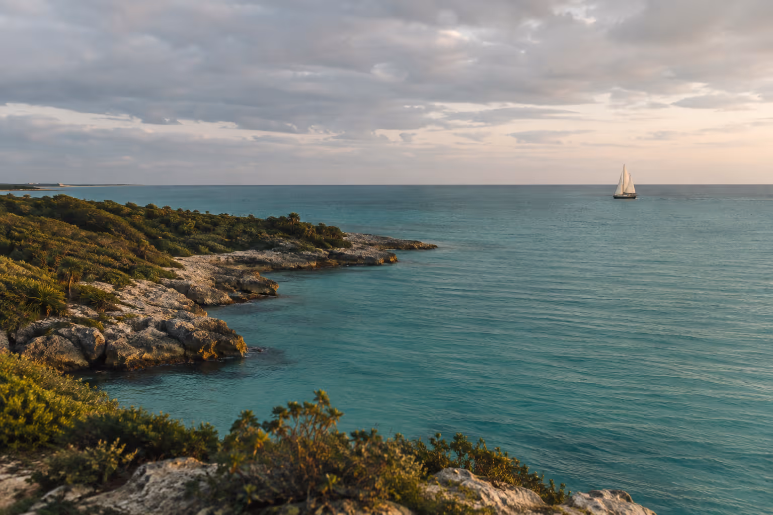 A sailboat crosses turquoise water beside a rocky, shrub-covered shoreline under a cloudy sky.