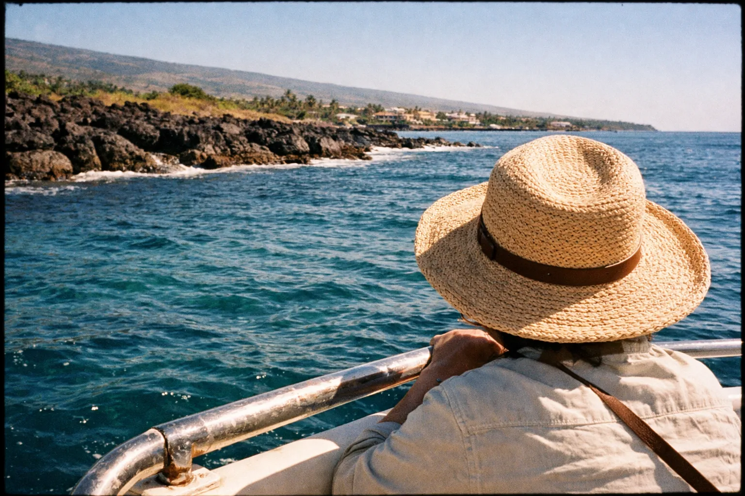 A person in a straw hat looks at the rocky Kona coastline from a boat on the ocean.