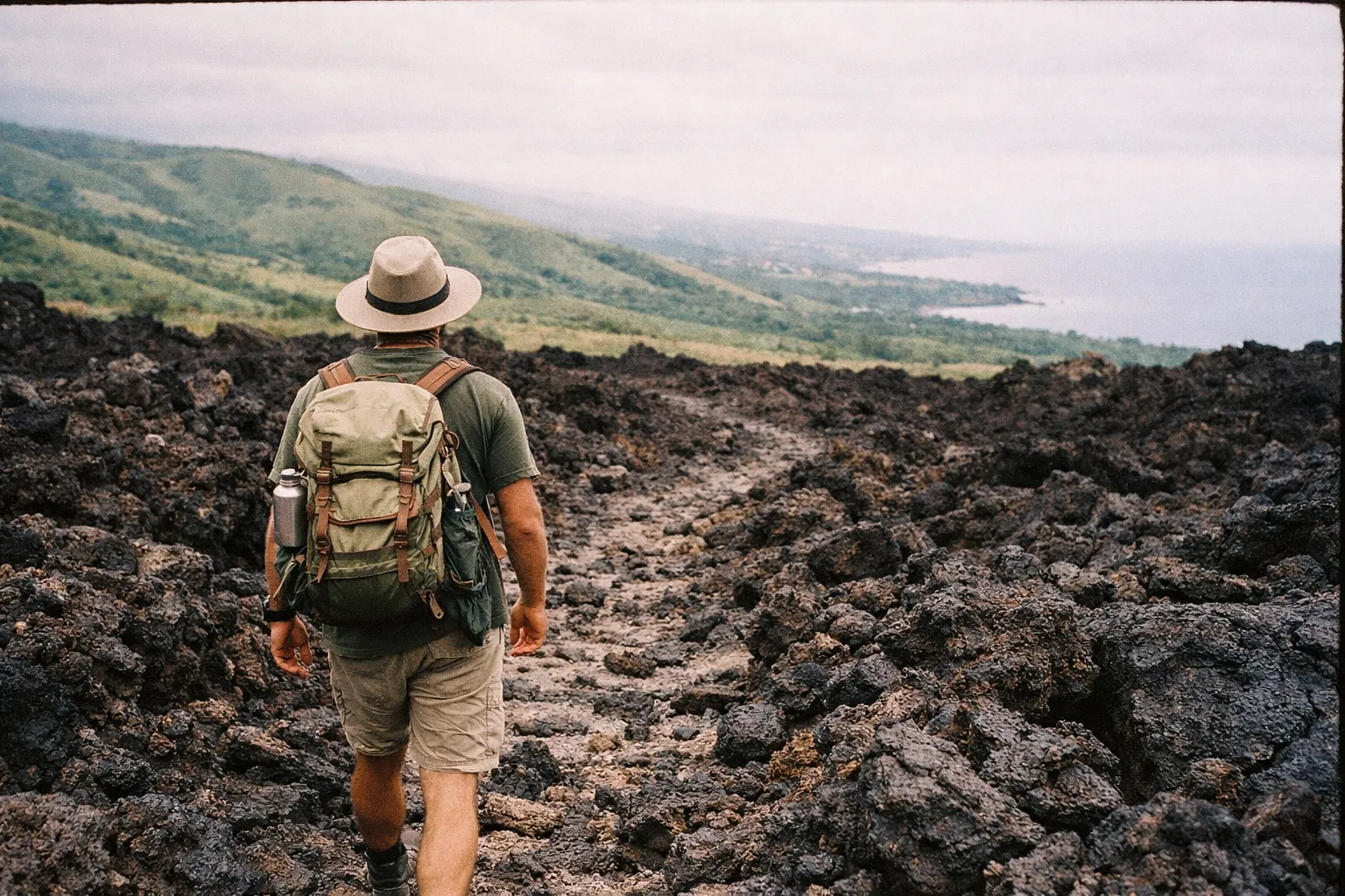 A hiker with a backpack walks along a lava field trail overlooking green hills and the ocean in Kona, Hawaii.