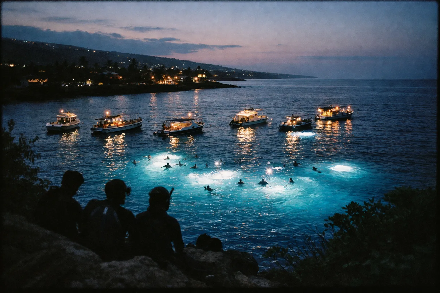 Night snorkelers with lights gather near boats in the waters off Kona's coastline at dusk.