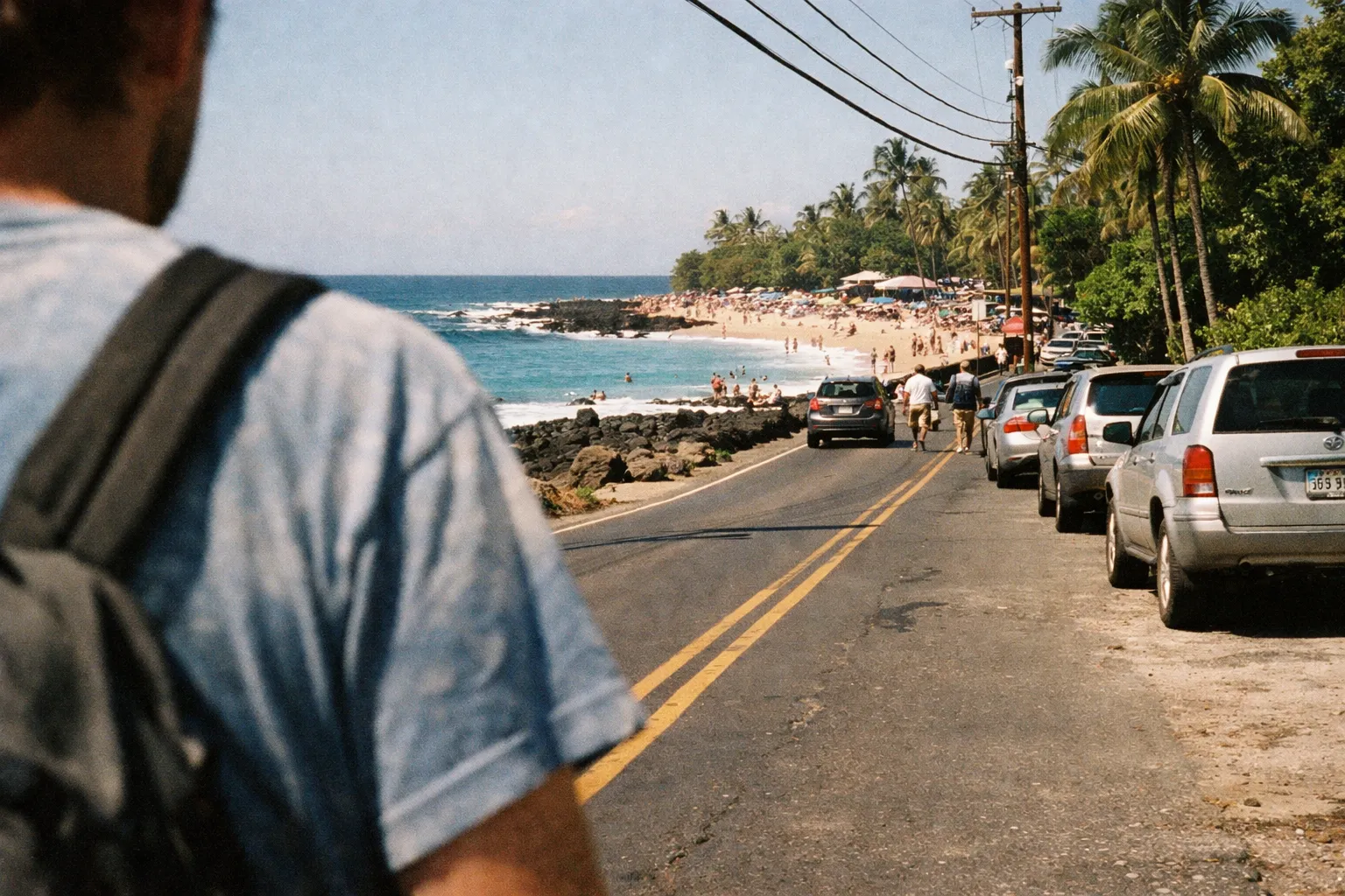A person walks beside a road lined with cars next to a busy beach and palm trees in Kona, Hawaii.