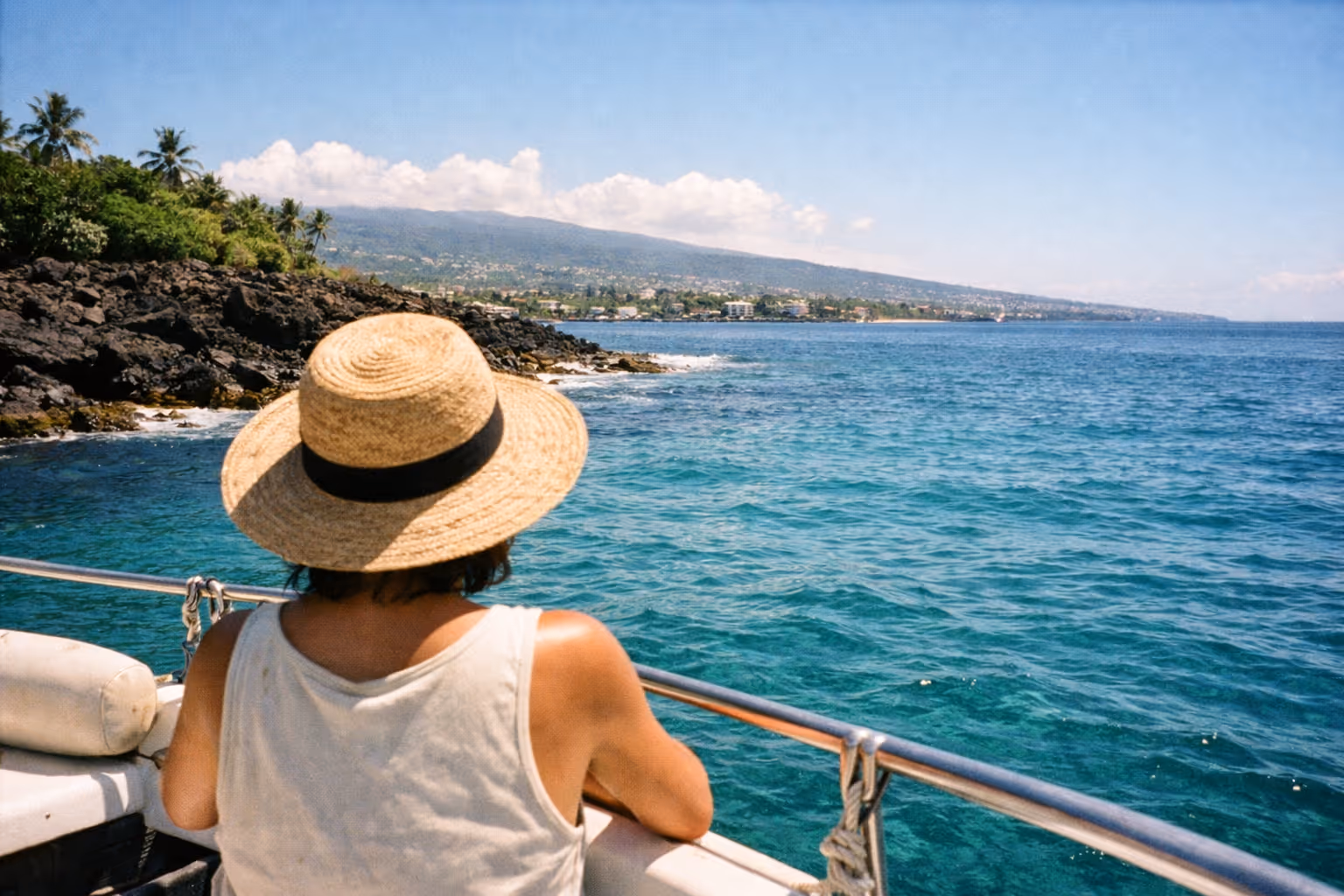 A person in a straw hat looks out at the Kona coastline from a boat on the ocean.