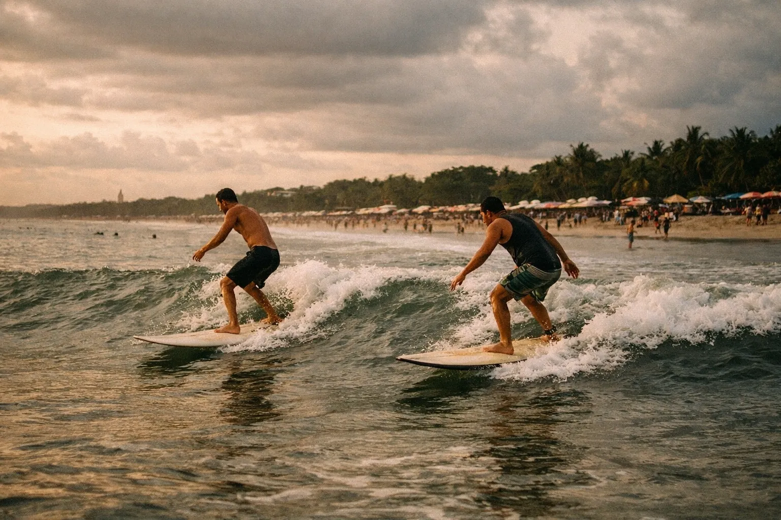 Two people surfing small waves near a crowded beach lined with palm trees and umbrellas.