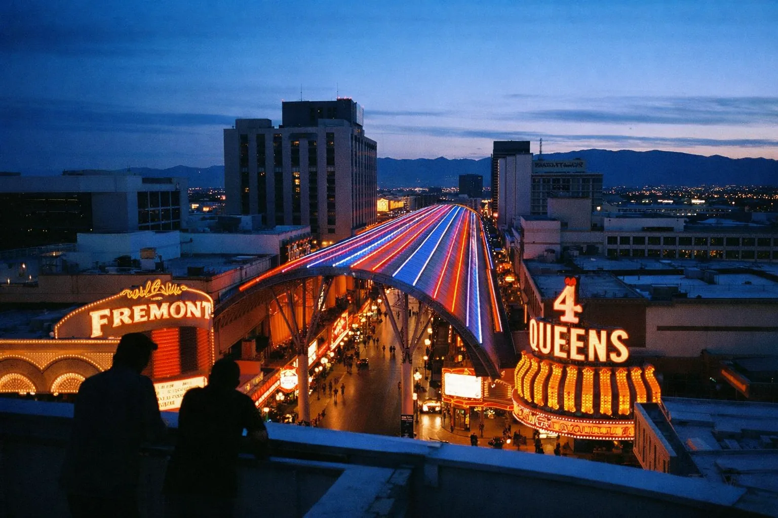 People overlook Fremont Street in downtown Las Vegas at dusk with neon lights and casinos visible.