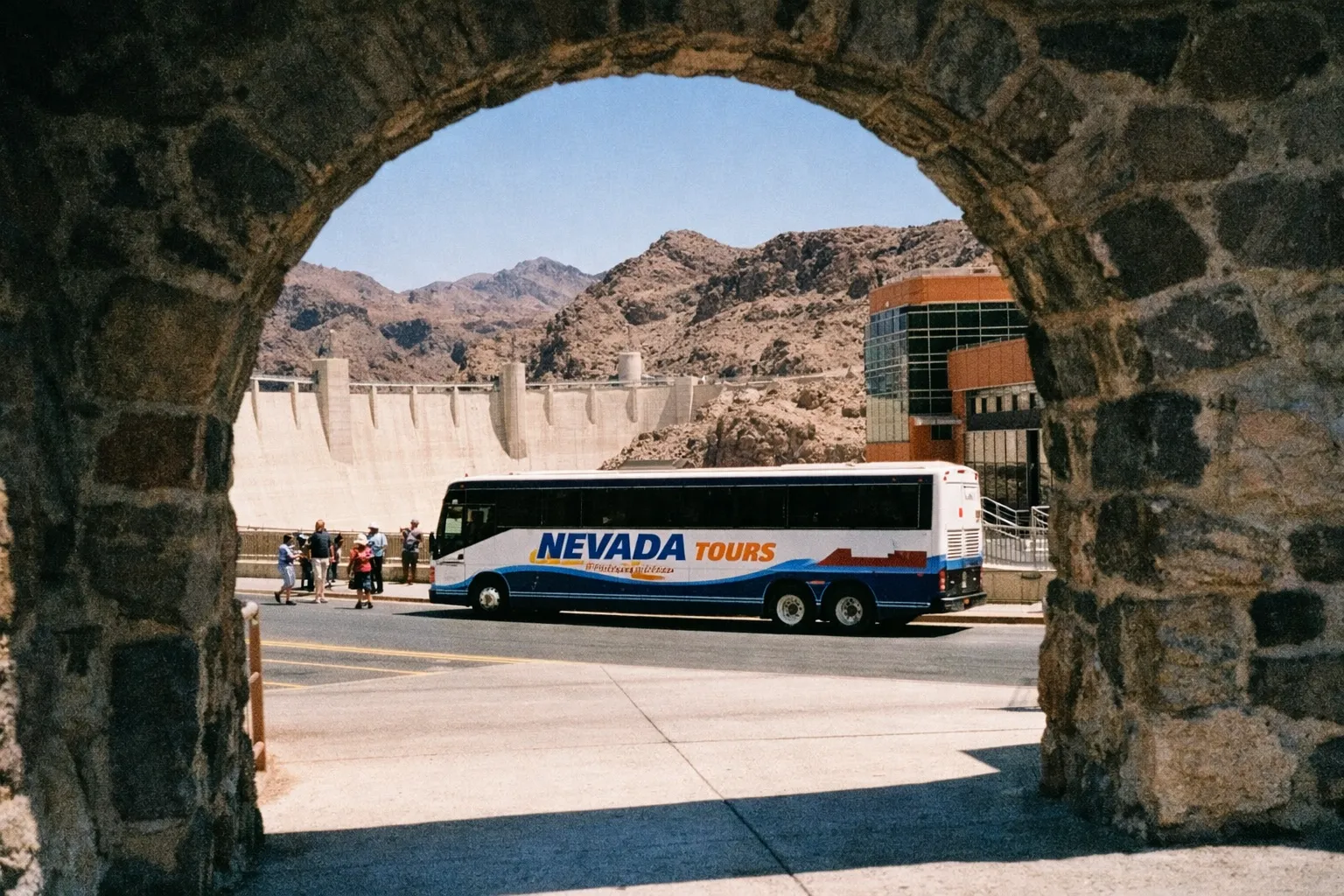 A tour bus and visitors stand near the Hoover Dam with rocky hills in the background.