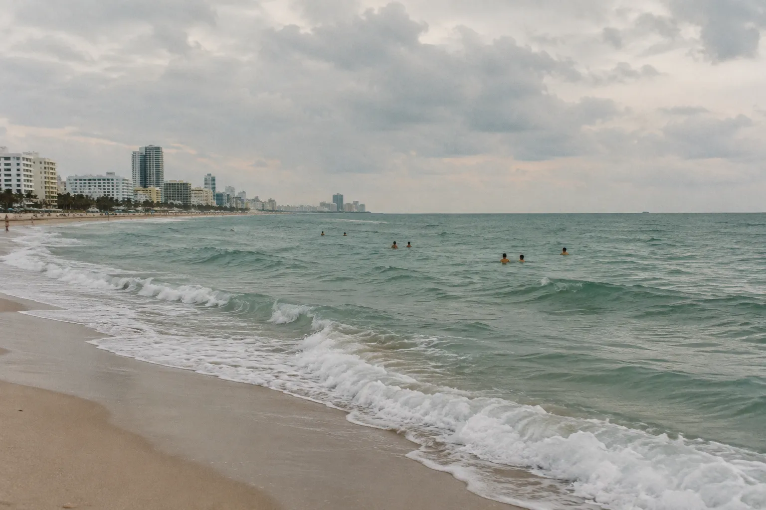Swimmers wade off Miami Beach beneath a cloudy sky and a line of coastal buildings.