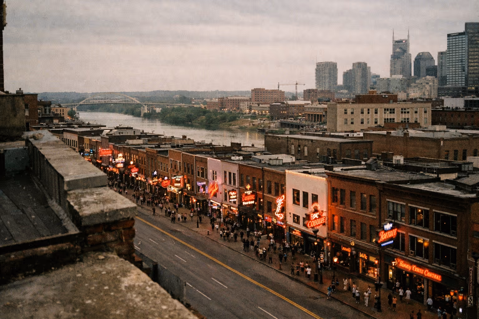 Crowds walk along Nashville's Broadway with neon-lit bars and honky tonks, the skyline and river in the background.