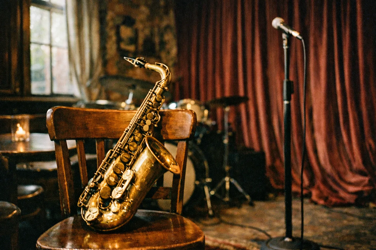 A saxophone rests on a wooden chair in a dimly lit jazz club with drum set and microphone on stage.
