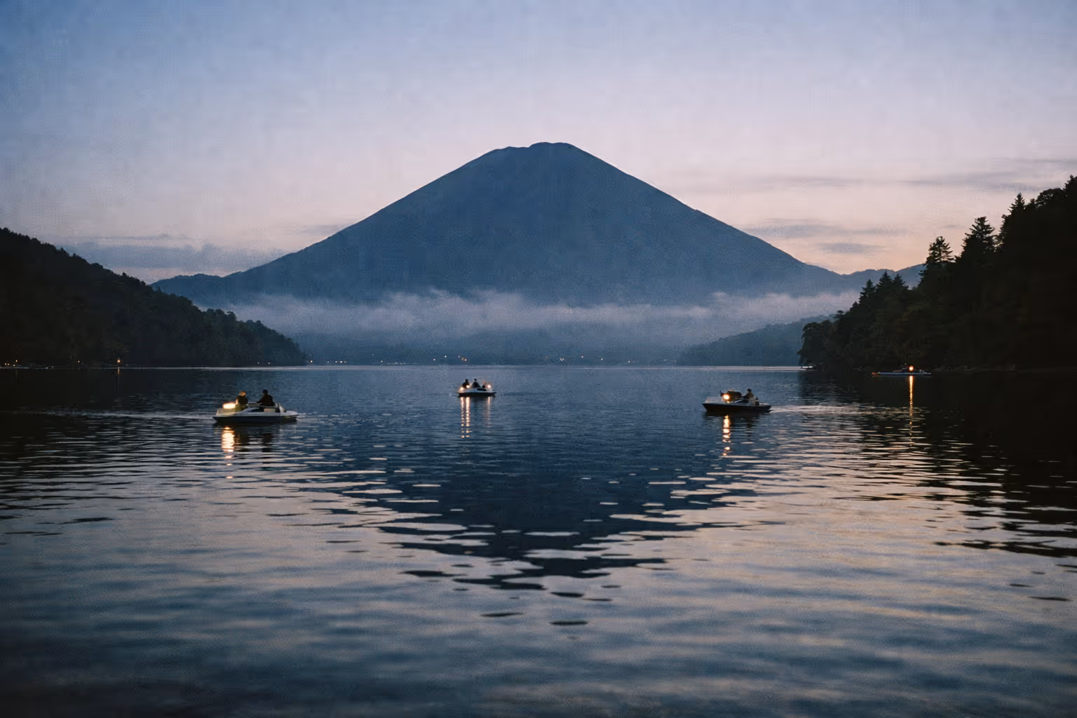 Small boats with people float on Lake Chuzenji at dusk with Mount Nantai in the background.