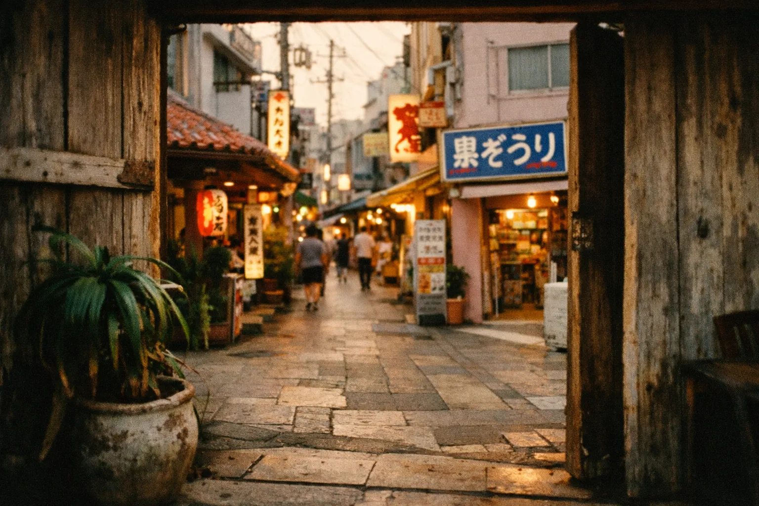 A narrow street in Okinawa lined with shops and glowing signs in the early evening.