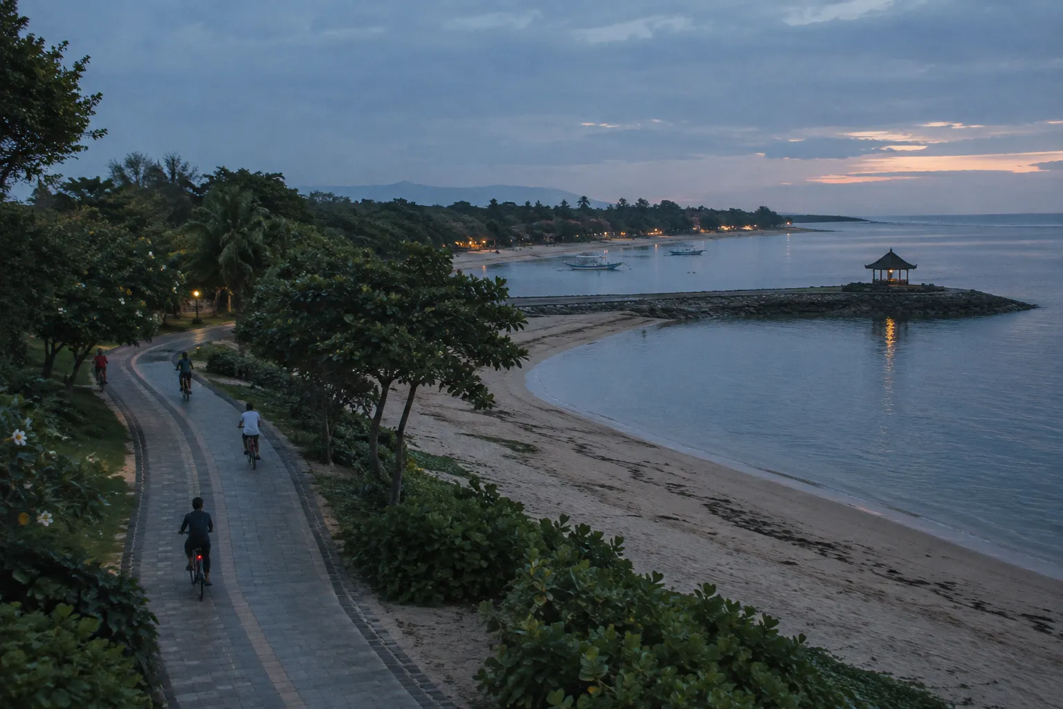 Cyclists ride along a palm-lined beach path beside calm water at sunset in Sanur, Bali.