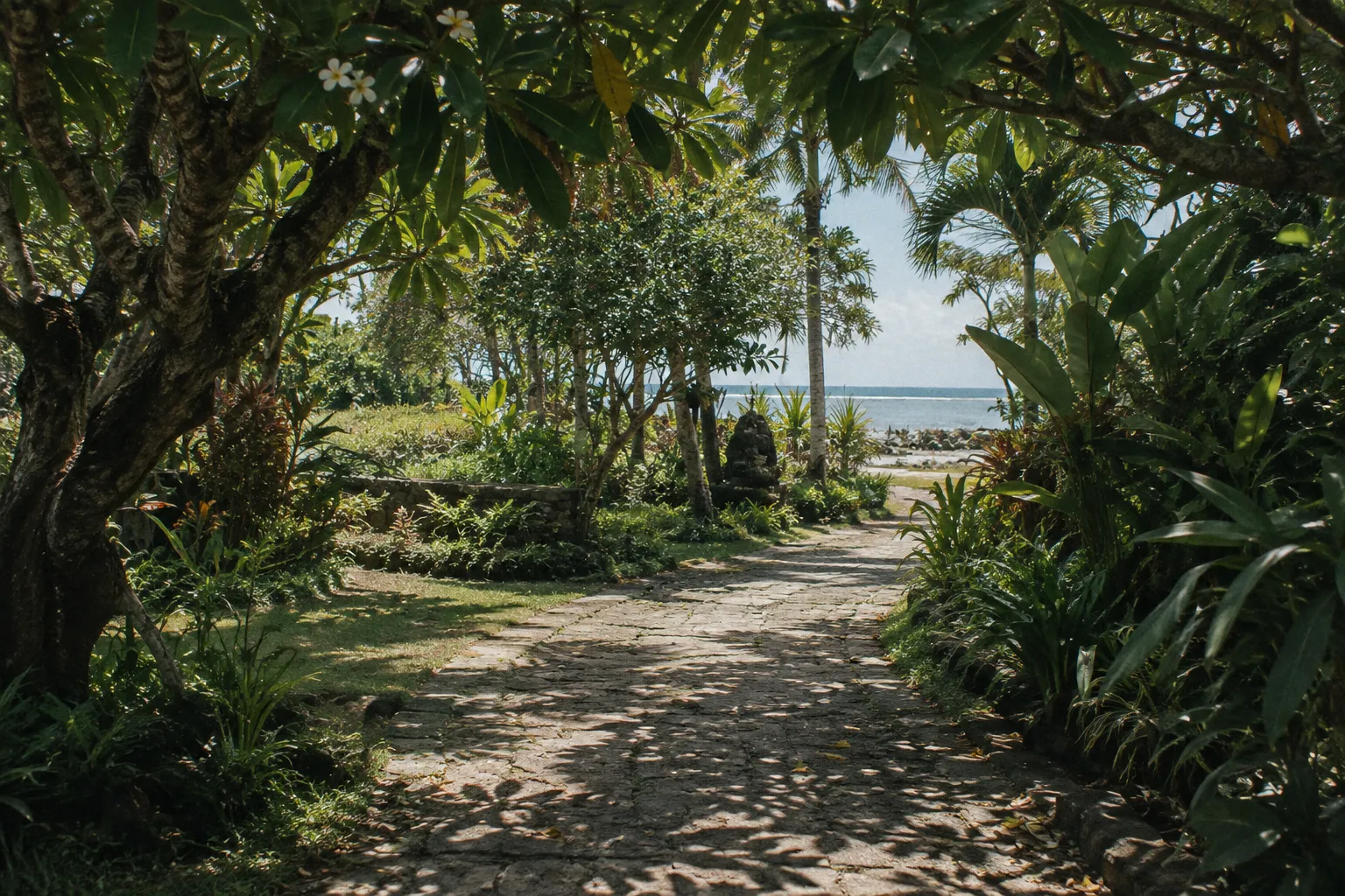 A shaded stone path winds through tropical gardens toward the beach in Sanur, Bali.