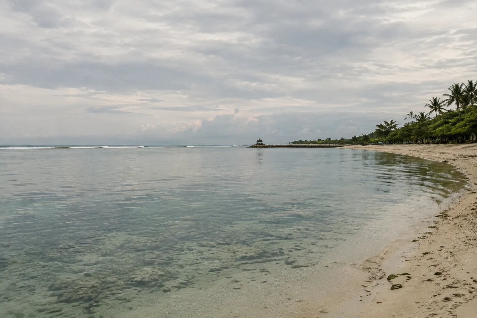 Calm shallow water curves along a sandy beach lined with palms in Sanur, Bali.