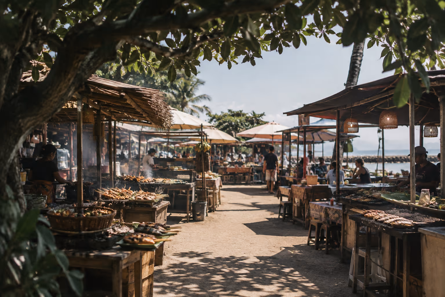 Open-air food stalls line a shaded path beside the beach in Sanur, Bali.